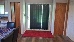 A room with a wooden floor, a red carpet, a dining table with a patterned tablecloth, and a window with green curtains. - 4-BEDROOM APARTMENT FOR ALL-INCLUSIVE WORKERS | 5 MN PONT LAVIOLETTE  (Trois-Rivières)