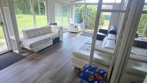 A spacious sunroom with large sliding glass doors, a white sofa, a chair, and a small table with a colorful cloth. - 4-BEDROOM APARTMENT FOR ALL-INCLUSIVE WORKERS | 5 MN PONT LAVIOLETTE  (Trois-Rivières)