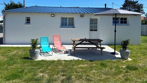 A small white house with a metal roof, a patio with two lounge chairs, a picnic table, and a large umbrella. - Atypical gîte Les Epinettes, 10 minutes from the beach in the Baie de Somme  (Cayeux-sur-Mer)