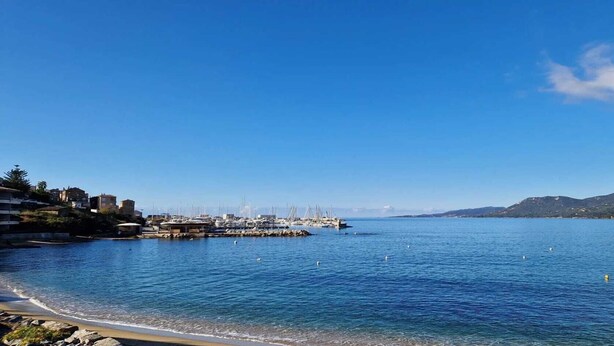 A coastal marina with boats docked, a clear blue sky, and a hilly landscape in the background.