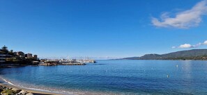 A coastal marina with boats docked, a clear blue sky, and a hilly landscape in the background.