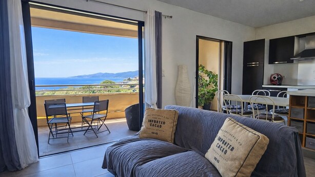 Ein modernes Wohnzimmer mit einer Couch, zwei Kissen, einem Essbereich mit Stühlen, einer Küche und einem Balkon mit Meerblick.