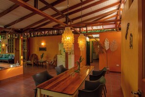 A spacious dining area with a wooden table and chairs, a hanging light fixture, and a view into another room.