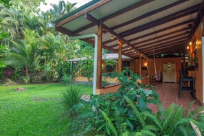 A covered outdoor area with a wooden ceiling and hanging lights, surrounded by lush greenery and a small building with a tiled roof.