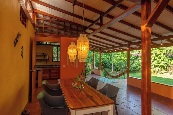 A dining area with a wooden table and chairs, a hanging light fixture, and a view of a hammock and greenery outside.