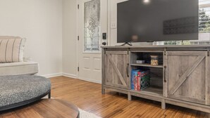 A living room with a sofa, ottoman, TV, and wooden cabinet with board games. - ModernNest-KS Bed| Fire Pit• Near TU•Expo•Downtown (Tulsa)