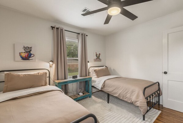 A bedroom with two single beds, a ceiling fan, a bedside table with a lamp, a window with curtains, and a wall-mounted artwork. - ModernNest-KS Bed| Fire Pit• Near TU•Expo•Downtown (Tulsa)