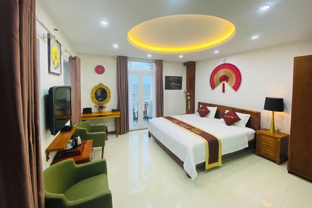 A hotel room with a bed, green chairs, a desk, a TV, and a view of the sea through the window.