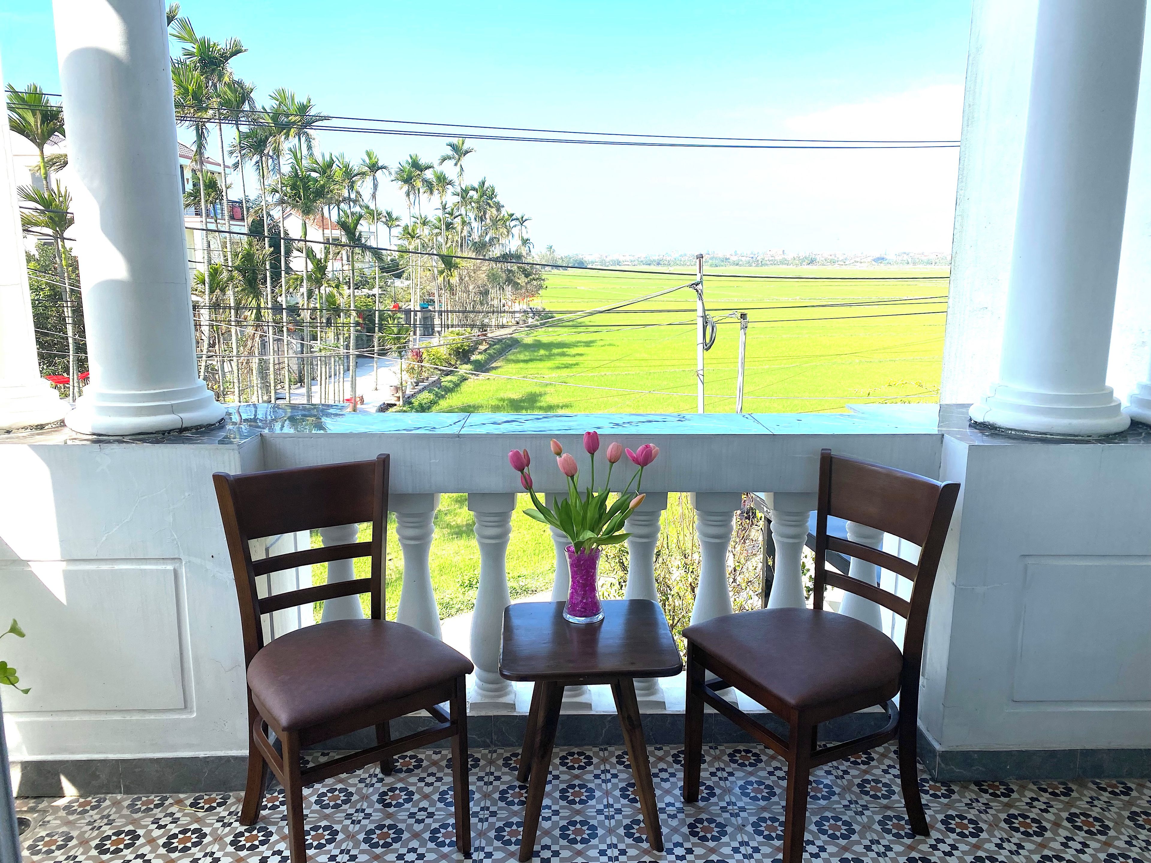 A balcony with two wooden chairs and a table, a vase of pink flowers, and a view of a green field and palm trees.