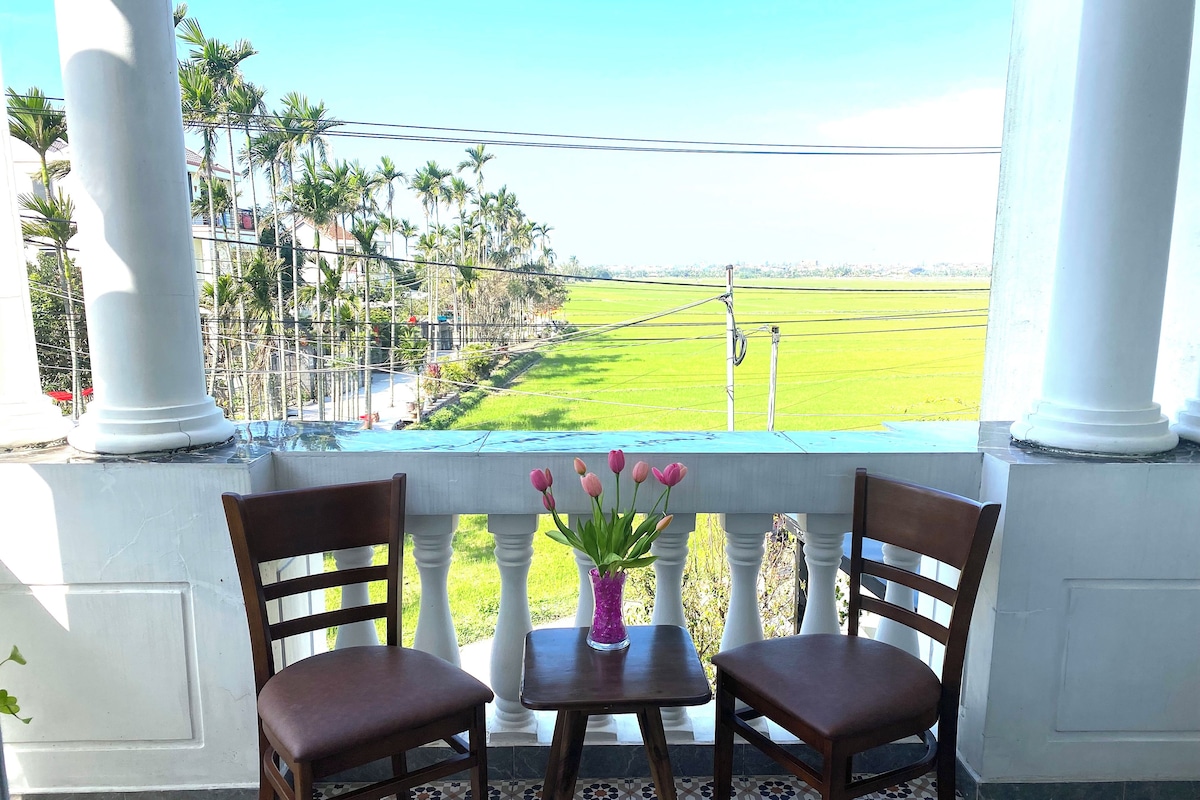 Un balcón con dos sillas de madera y una mesa, un jarrón con flores rosas y vistas a un campo verde y palmeras.