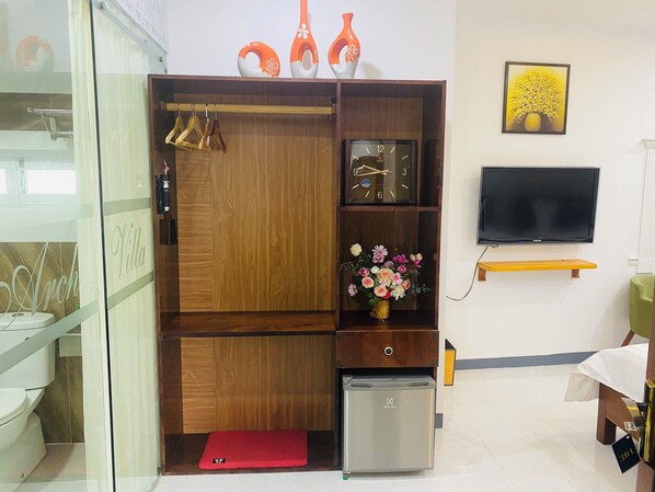 Habitación de hotel con armario de madera, refrigerador pequeño, televisor montado en la pared y un arreglo floral sobre la cómoda.