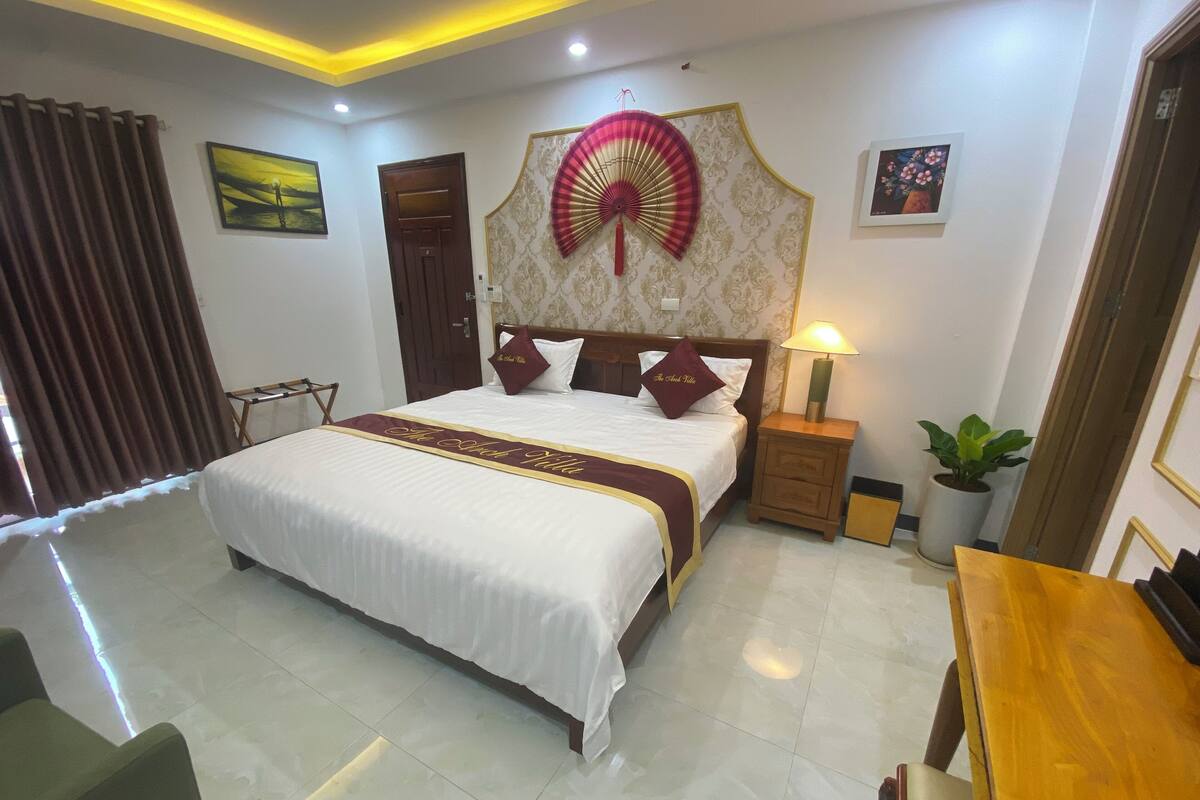 A hotel room with a large bed, a bedside table with a lamp, a potted plant, and a wooden chair.