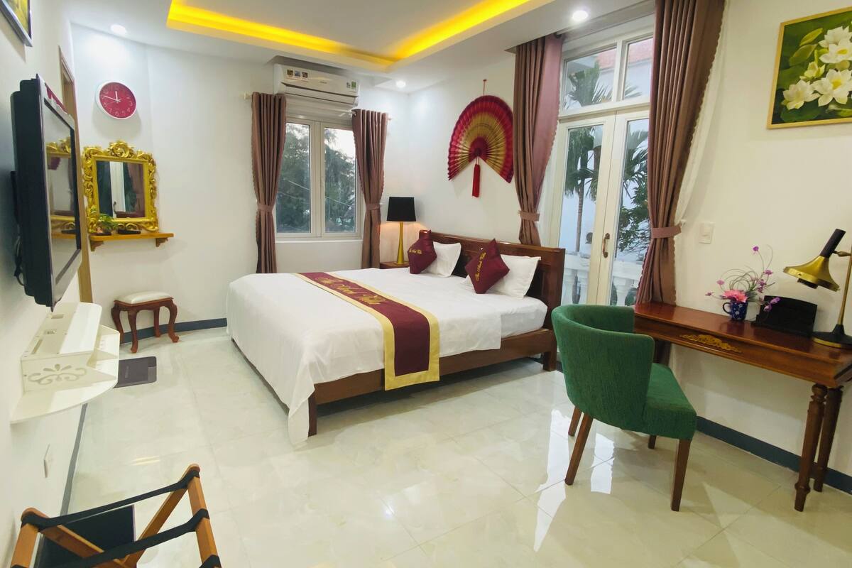 A hotel room with a bed, a desk, a chair, a mirror, a window, and an air conditioning unit.