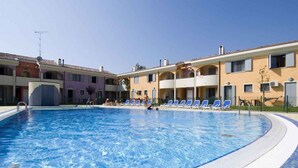 A swimming pool with lounge chairs, a building with balconies, and a clear sky. - Holiday apartment with air conditioning and garden (Bibione)