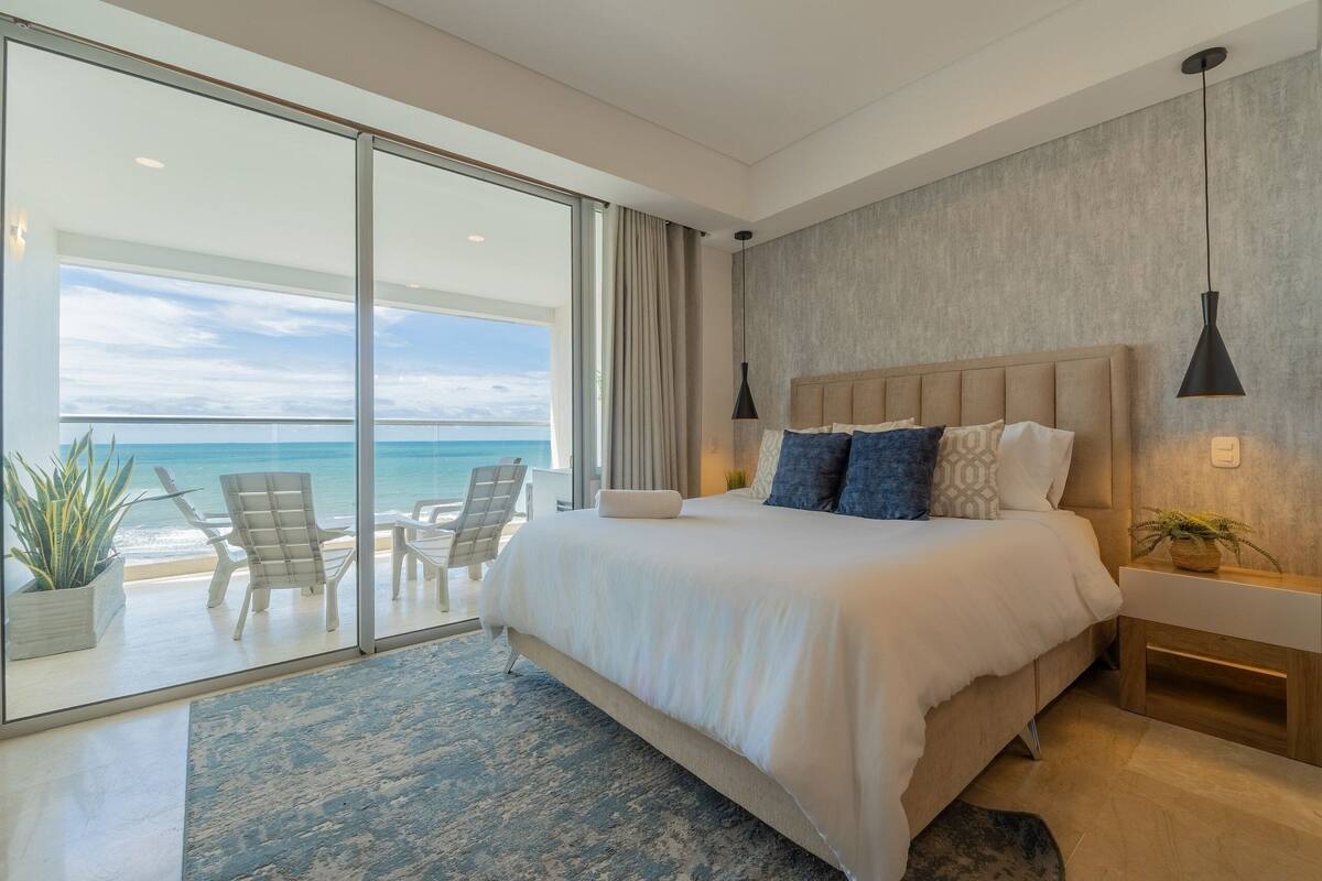 A modern bedroom with a large bed, a view of the ocean, and a balcony with seating.