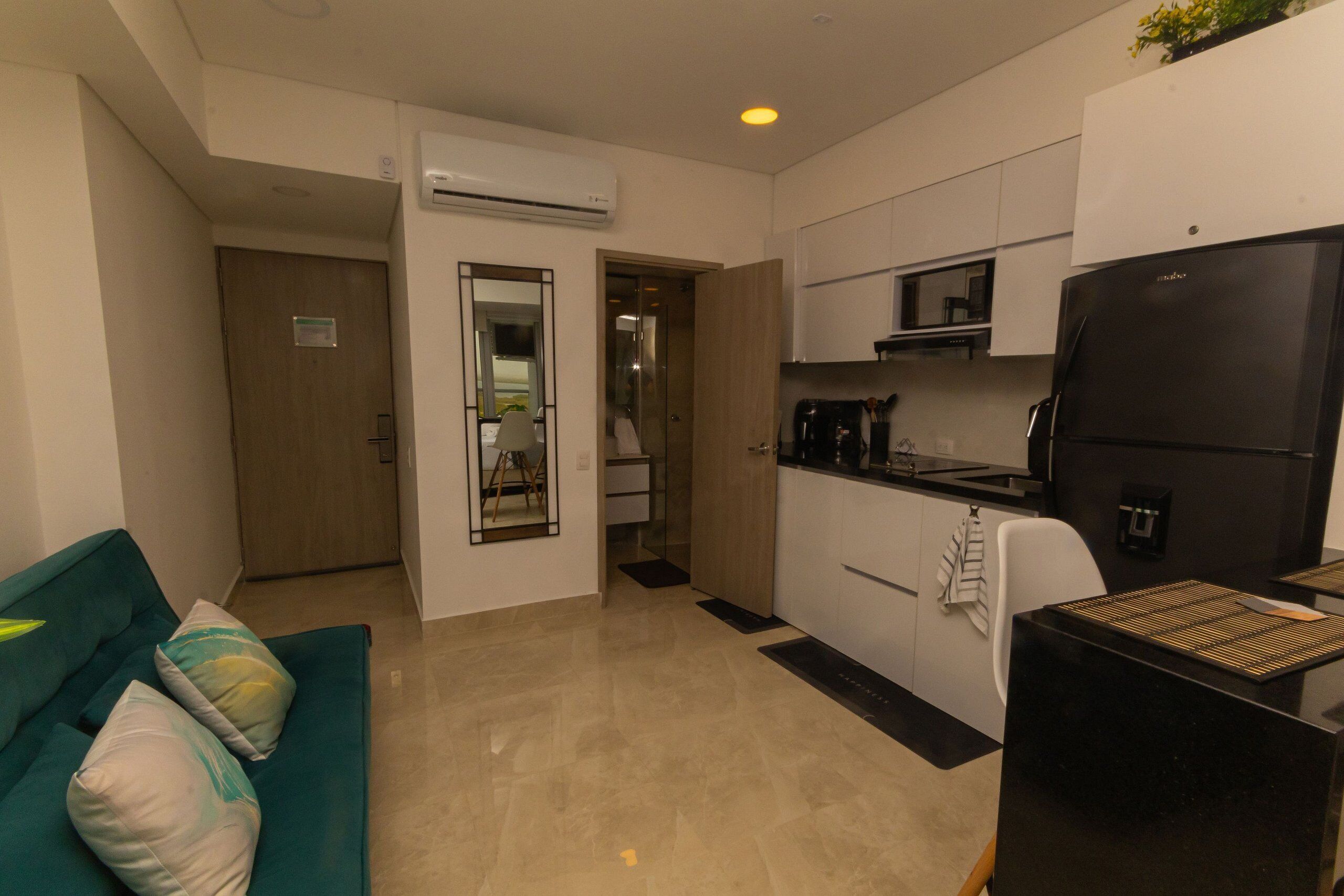 A modern kitchen with a black refrigerator, white cabinets, and a microwave. There is a teal sofa with cushions in the living area.
