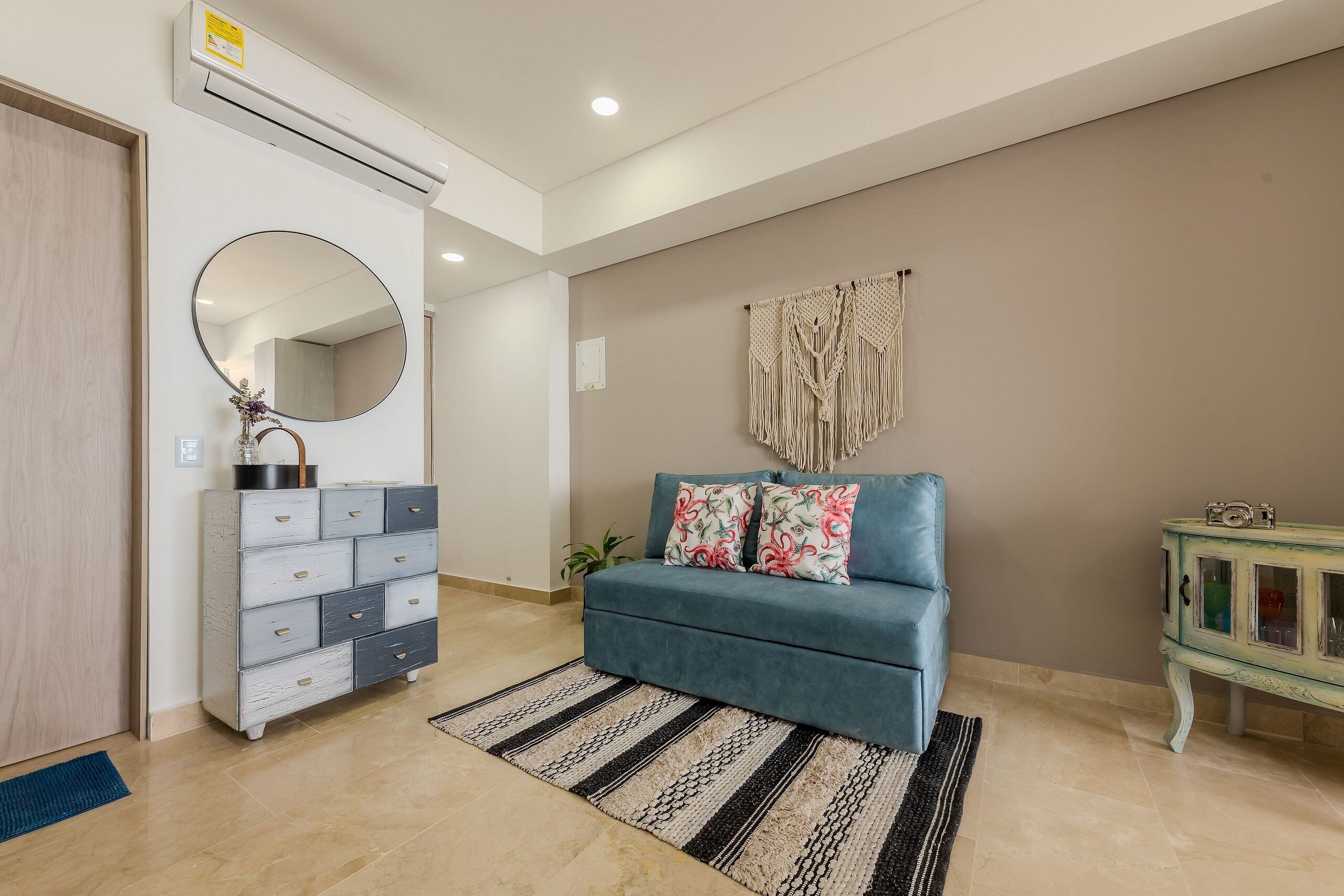 A living room with a blue sofa, a round mirror, a cabinet, and a macrame wall hanging.