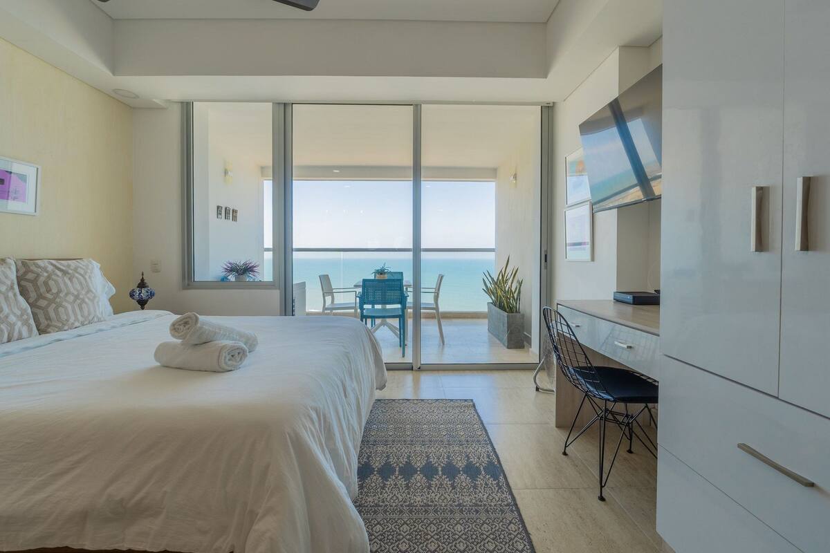 A modern hotel room with a large bed, a desk, and a view of the ocean through a sliding glass door.