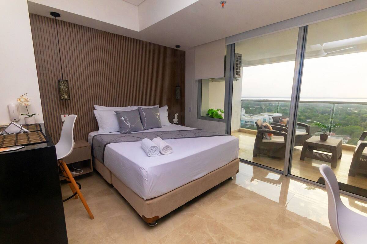 A modern bedroom with a large bed, a desk, and a balcony with outdoor seating.