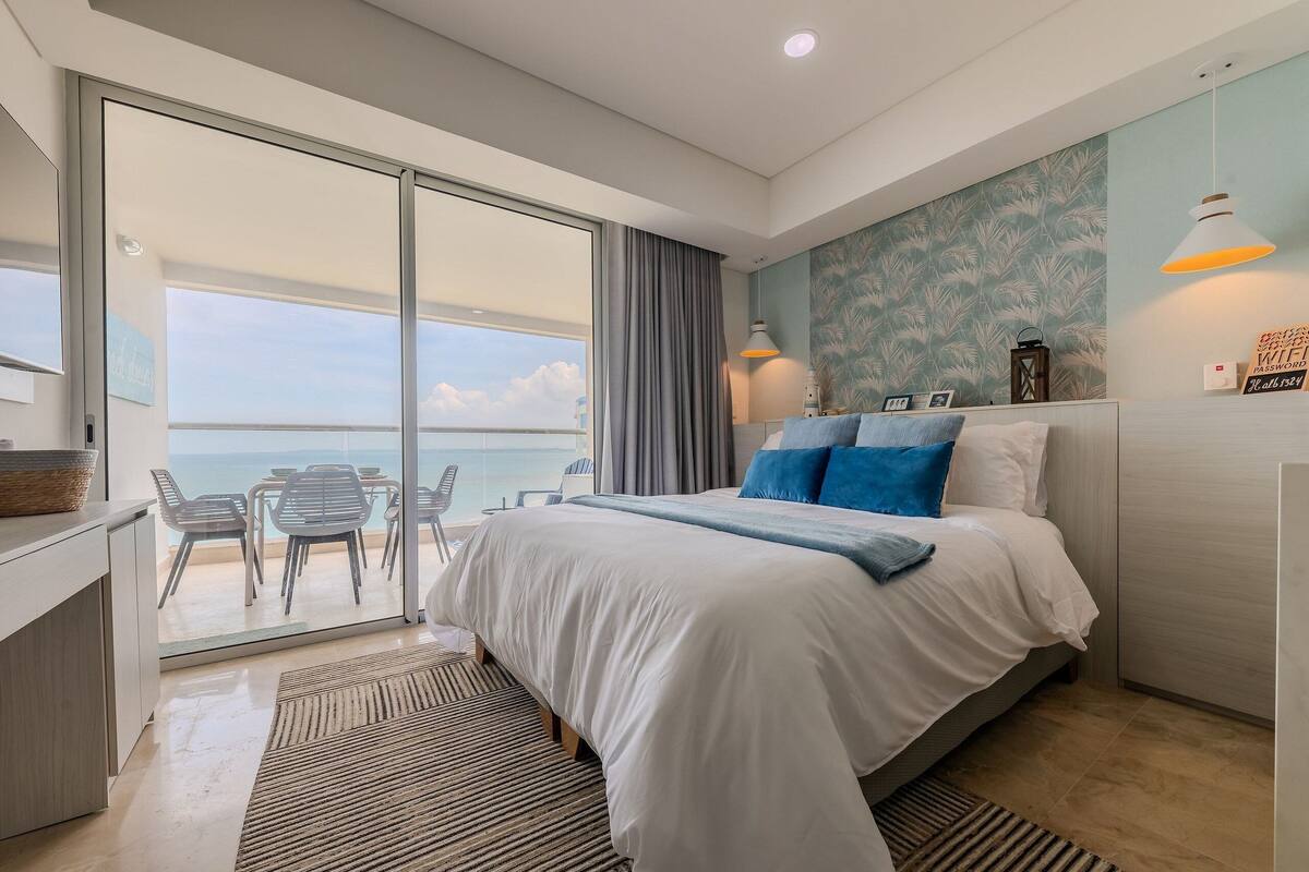 A modern hotel room with a large bed, a balcony with a view, and a dining area with chairs.