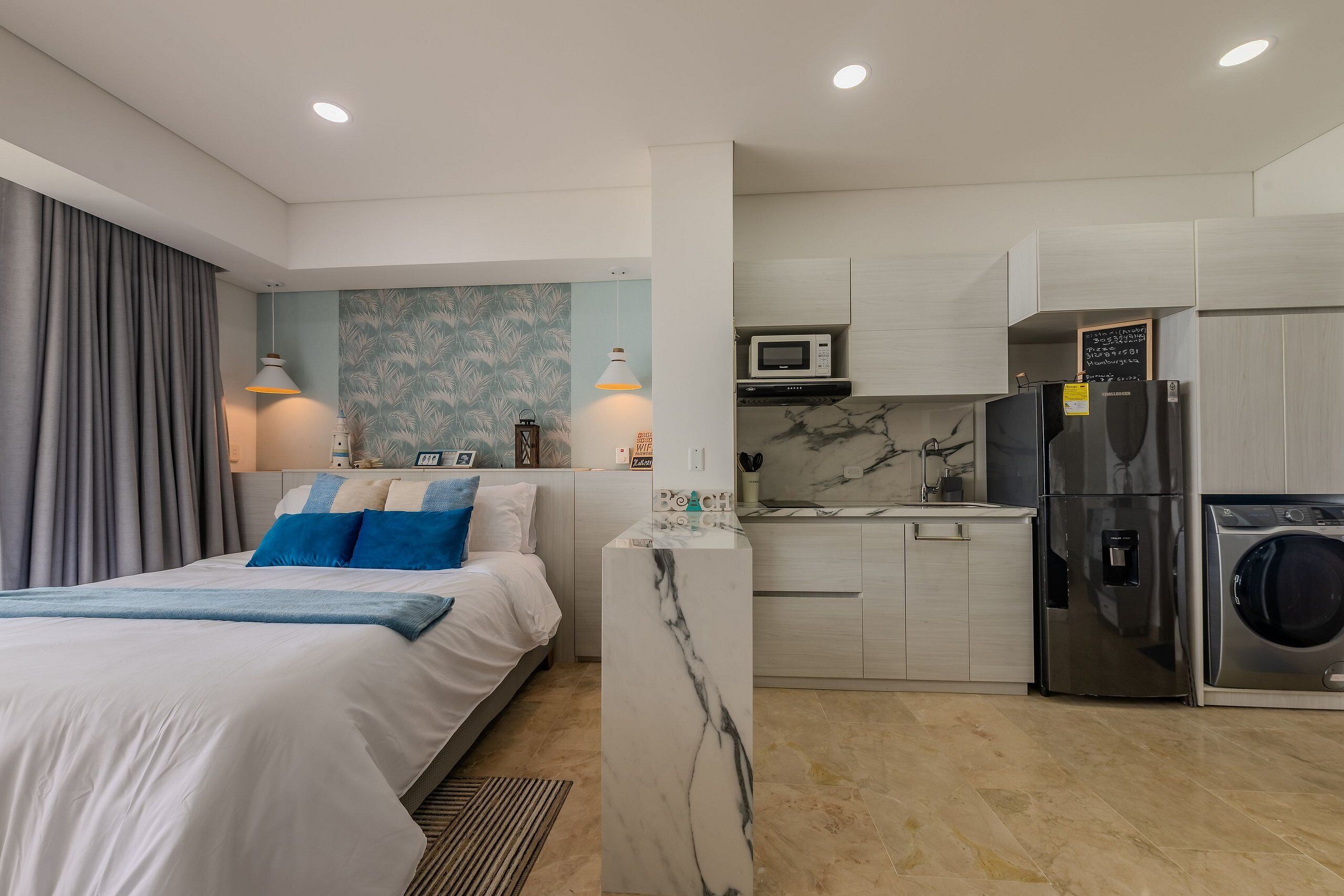 A modern hotel room with a bed, a marble accent wall, a kitchen area with a microwave, refrigerator, and washing machine.