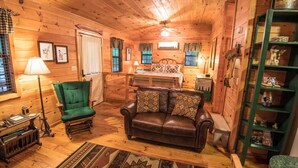 Ein gemütliches Blockhaus-Interieur mit Holzboden, einem grünen Sessel, einer Ledersofa und einem Bett, sowie einer grünen Regaleinheit.