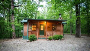 Eine Blockhütte mit Veranda, Schornstein und einer Schotterauffahrt, umgeben von Bäumen.