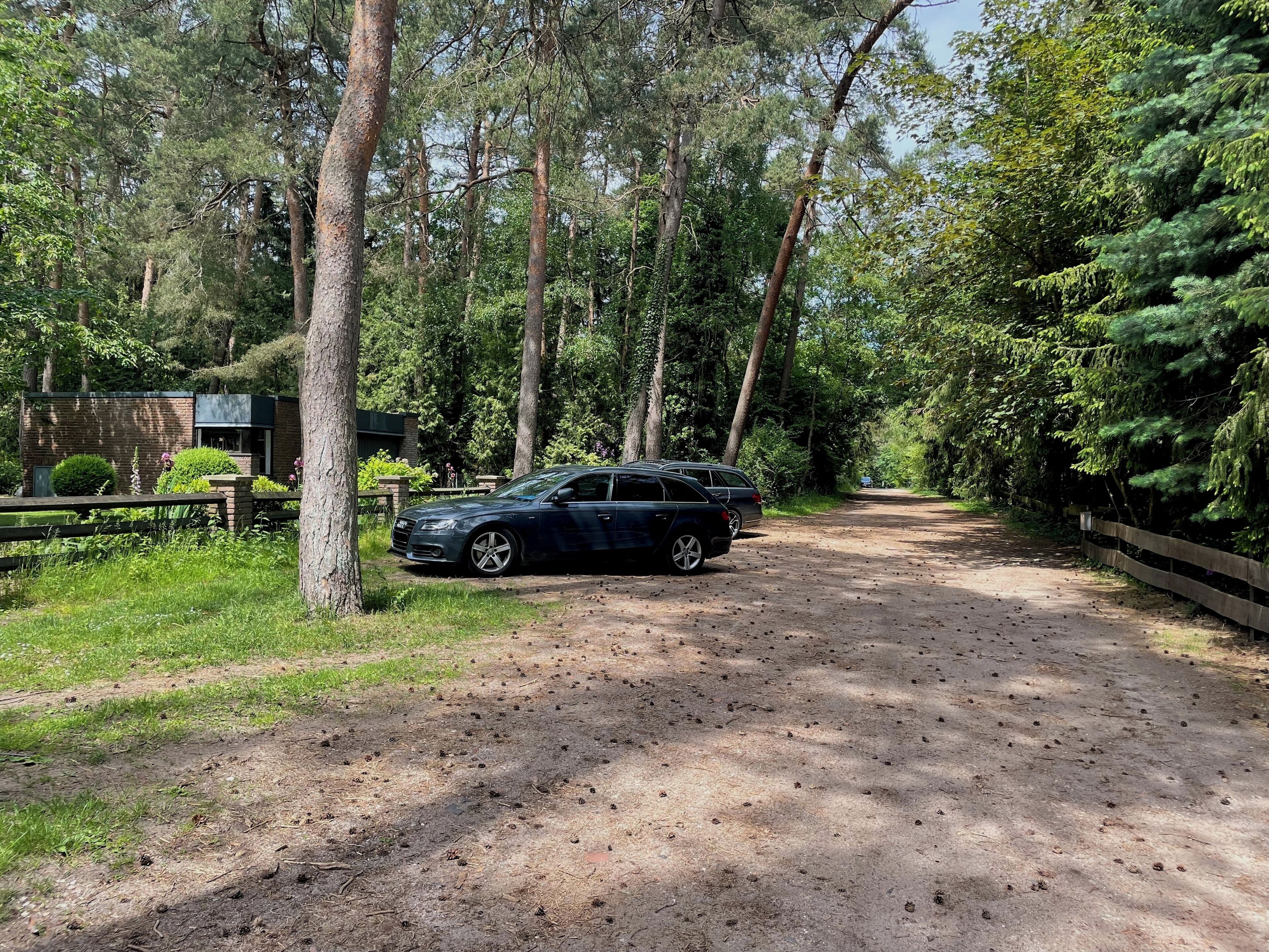 Ein unbefestigter Weg führt durch bewaldetes Gelände, an dem ein geparktes Auto und ein Backsteinhaus mit grüner Dachbegrünung stehen.