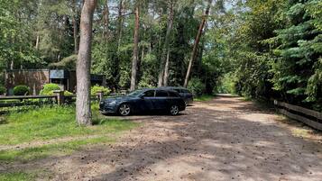 Ein unbefestigter Weg führt durch bewaldetes Gelände, an dem ein geparktes Auto und ein Backsteinhaus mit grüner Dachbegrünung stehen.