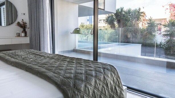 Ein modernes Schlafzimmer mit einem großen Bett, einem Nachttisch mit einer Vase und Blick auf einen Balkon mit Pflanzen und Stadtansicht.