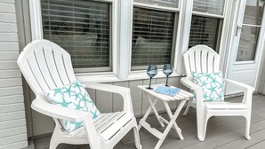 Dos sillas Adirondack blancas con cojines estampados de estrellas de mar, una mesita blanca con dos copas de vino y un libro, y una ventana con persianas.