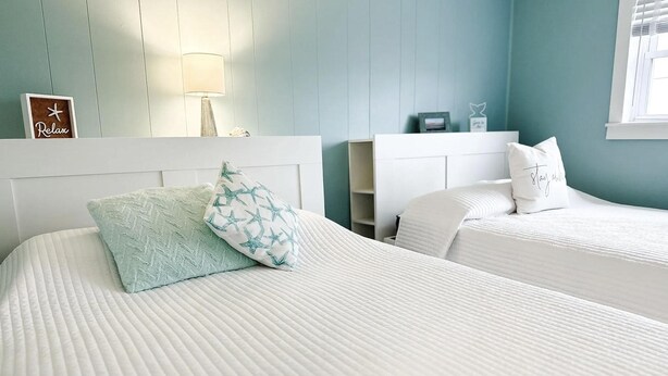 Habitación de hotel con dos camas, cada una con cabecero blanco y pared azul claro. Hay una lámpara a la izquierda de la habitación y un pequeño estante con objetos decorativos.