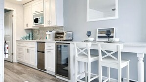 Una cocina moderna con armarios blancos, un microondas integrado, un tostador y un lavavajillas. Hay una barra con sillas blancas y una copa de vino.