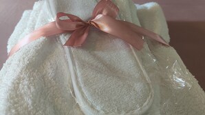 Une serviette soigneusement pliée, ornée d’un ruban rose, est posée sur un lit dont la tête de lit est rose et qui est agrémenté d’un oreiller blanc.