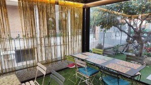 Un coin repas avec une table dressée pour quatre personnes, entourée de chaises dotées de coussins. On y trouve un rideau décoratif et, par la fenêtre, on peut admirer un arbre et une clôture.
