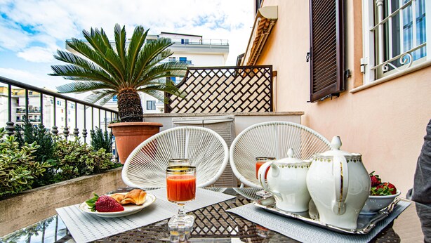 Un balcon avec une table préparée pour le petit-déjeuner, comprenant une assiette de nourriture, un verre de jus et des pichets en céramique blanche.