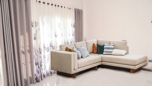 A modern living room with a beige sectional sofa, multiple pillows, and a large window with sheer curtains. - RM Homestay (Negombo)