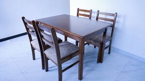 A rectangular wooden dining table with four chairs, each with cushioned seats and backs. The table is positioned against a white wall with a tiled floor. - RM Homestay (Negombo)