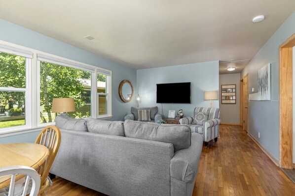 A living room with a grey sofa, a TV mounted on the wall, and a dining area with a wooden table and chairs.