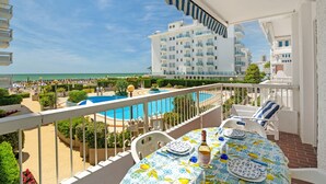 A balcony with a pool view, white furniture, and a table with a patterned tablecloth.