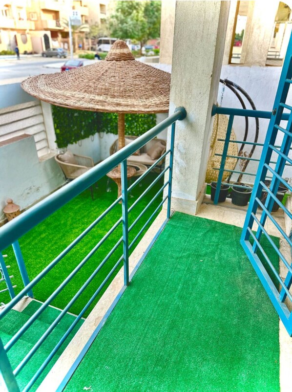 A balcony with artificial green turf, a metal railing, a wicker umbrella, and potted plants. - The Privacy Villa (6th of October City)