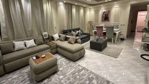 A modern living room with a sectional sofa, ottoman, and a small coffee table. There is a dining area with a table and chairs, and a painting on the wall. - The Privacy Villa (6th of October City)