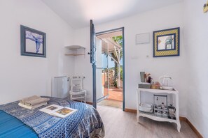 A bedroom with a bed, a bedside table with a kettle, a framed picture, and a door leading to an outdoor area.
