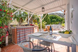 An outdoor patio with a table set for dining, a grill, and a view of the surrounding garden and hills.