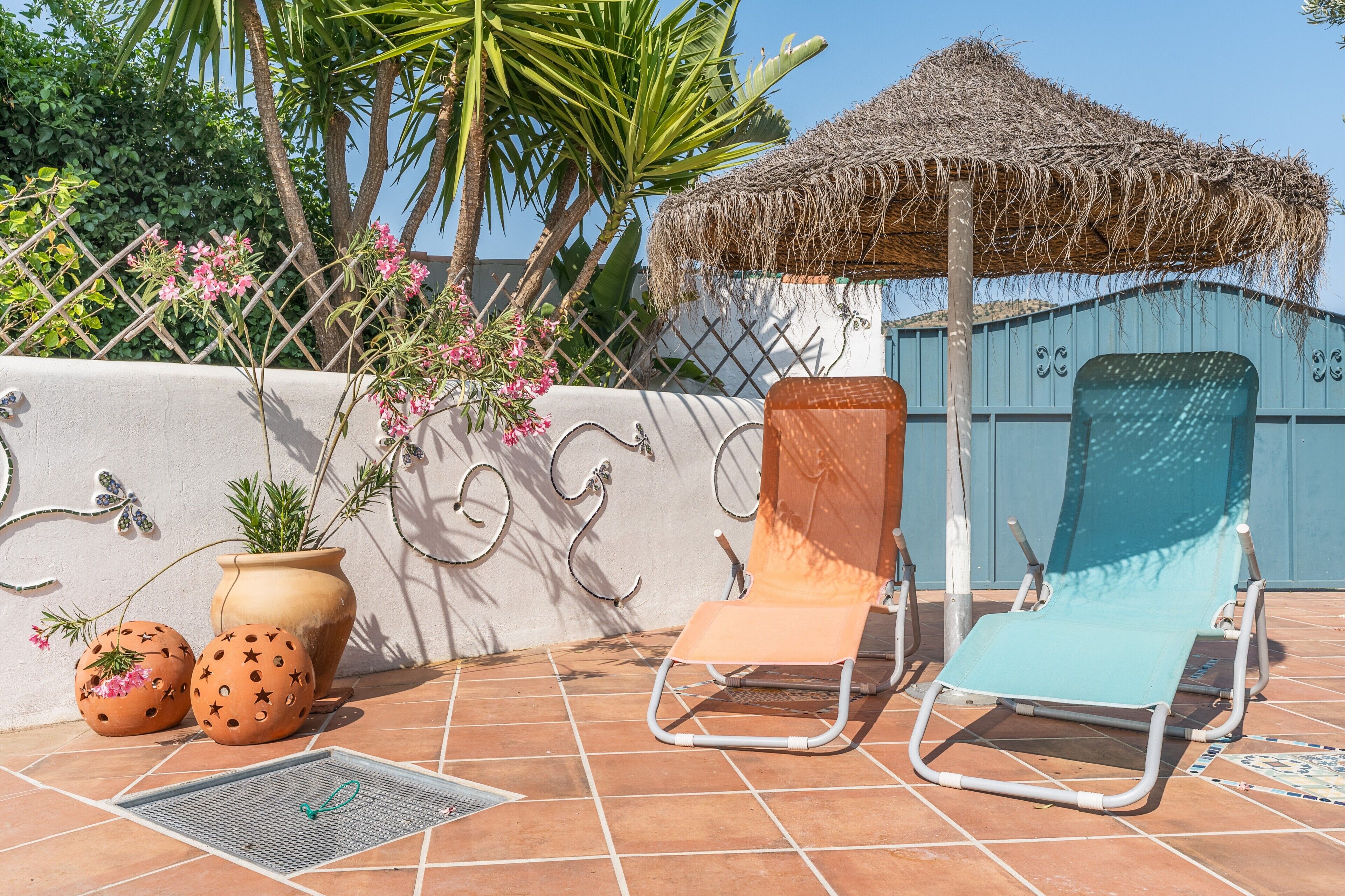 A terrace with two lounge chairs, a thatched umbrella, potted plants, and a decorative wall.