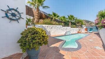 Una piscina con solárium de baldosas, un muro decorativo con timón de barco y plantas en macetas.