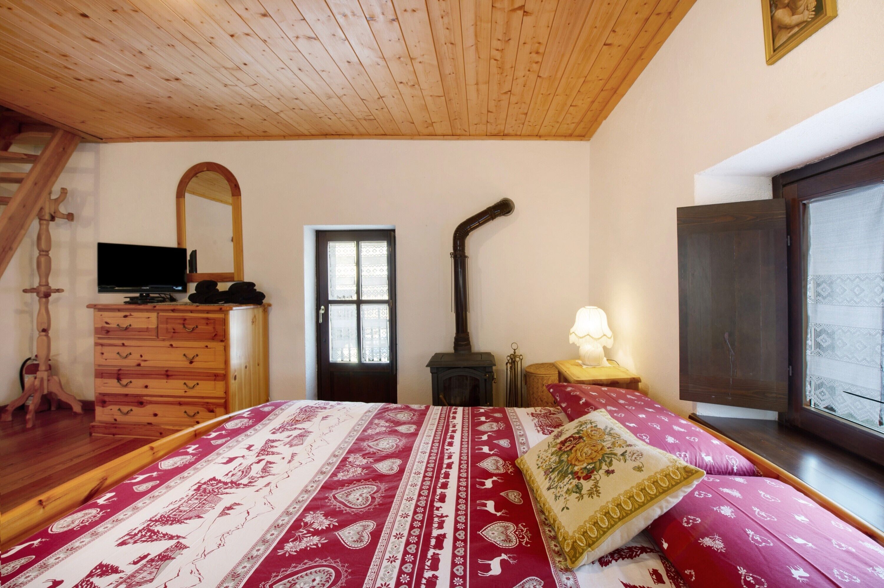 A bedroom with a wooden ceiling, a bed with patterned bedding, a dresser, a television, a fireplace, and a window.