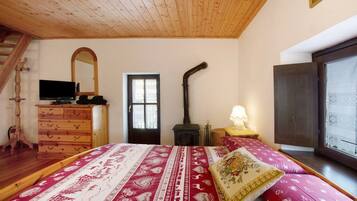 A bedroom with a wooden ceiling, a bed with patterned bedding, a dresser, a television, a fireplace, and a window.