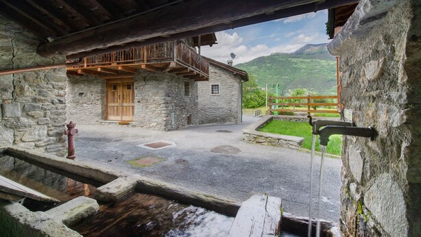 Een landelijk stenen gebouw met een houten balkon, een stenen waterput en uitzicht op een bergachtig landschap.