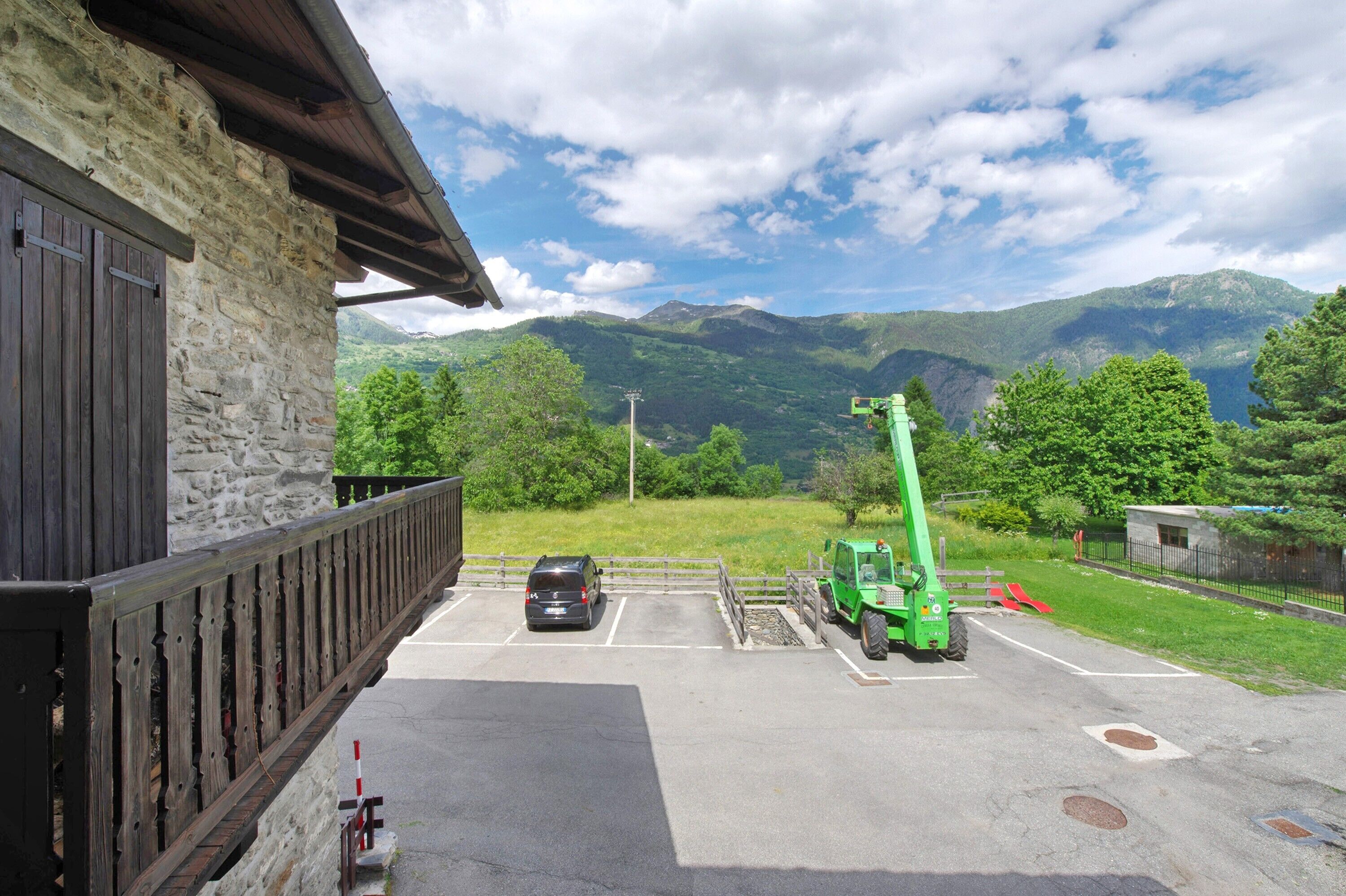 Ein Steinhaus mit Balkon, ein Parkplatz mit einem Auto und einem grünen Traktor, und eine gebirgige Landschaft im Hintergrund.